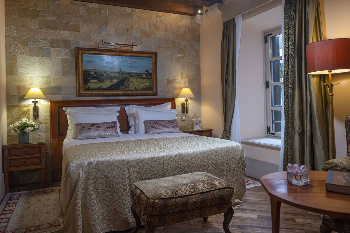 A hotel room with a large bed, two bedside tables with lamps, a small table with a book, a window with curtains, and a painting on the wall.