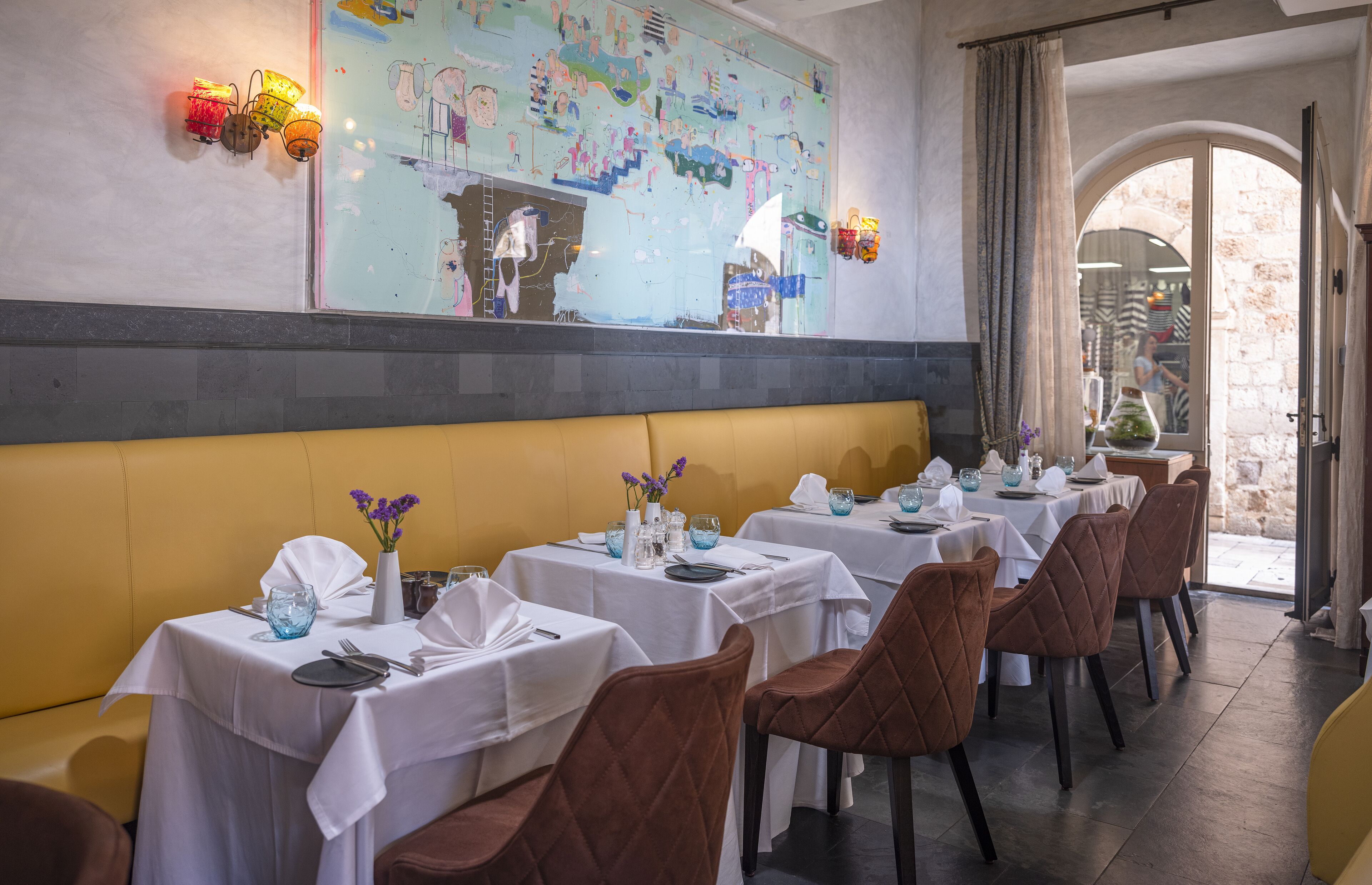 A restaurant with tables set for dining, a large decorative wall, and a view through an arched doorway.