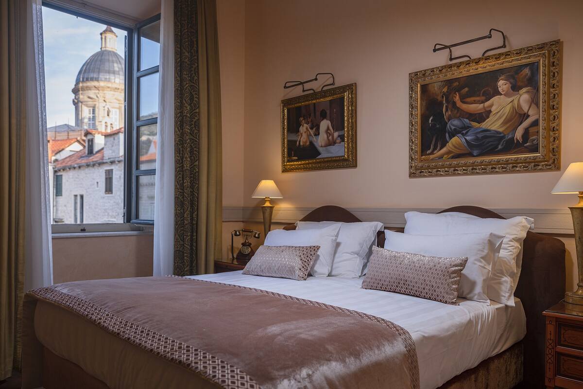 A hotel room with a large bed, two paintings on the wall, a window with a view of a building, and a nightstand with a lamp.