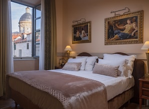A hotel room with a large bed, two paintings on the wall, a window with a view of a building, and a nightstand with a lamp.