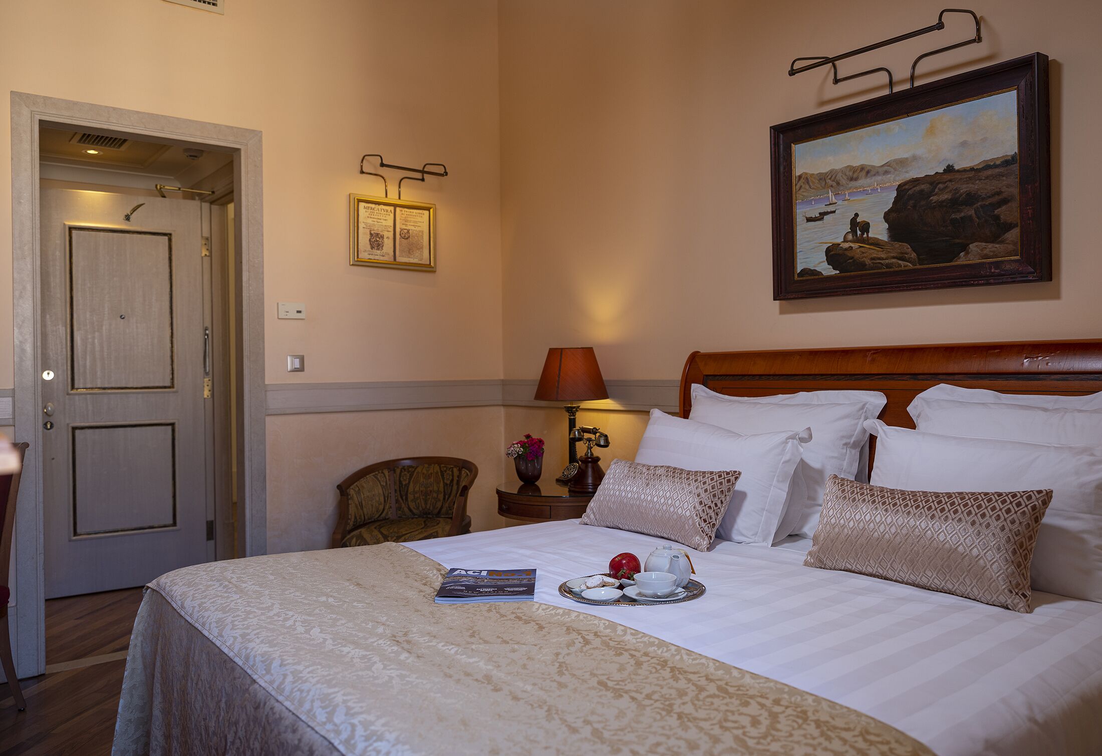 A neatly made bed with pillows, a bedside table with a lamp, a framed painting on the wall, and a door to the left.