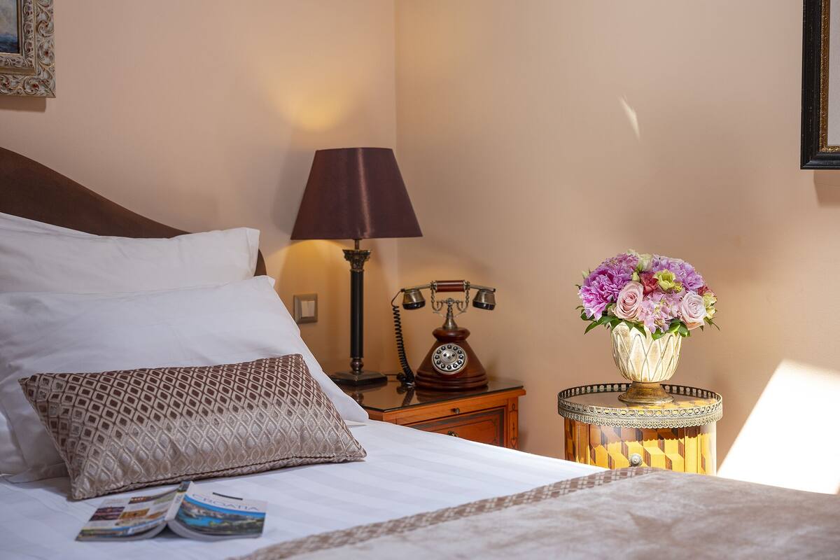 A neatly made bed with white linens, a patterned pillow, a vintage rotary phone, a lamp, and a vase with flowers on a bedside table.
