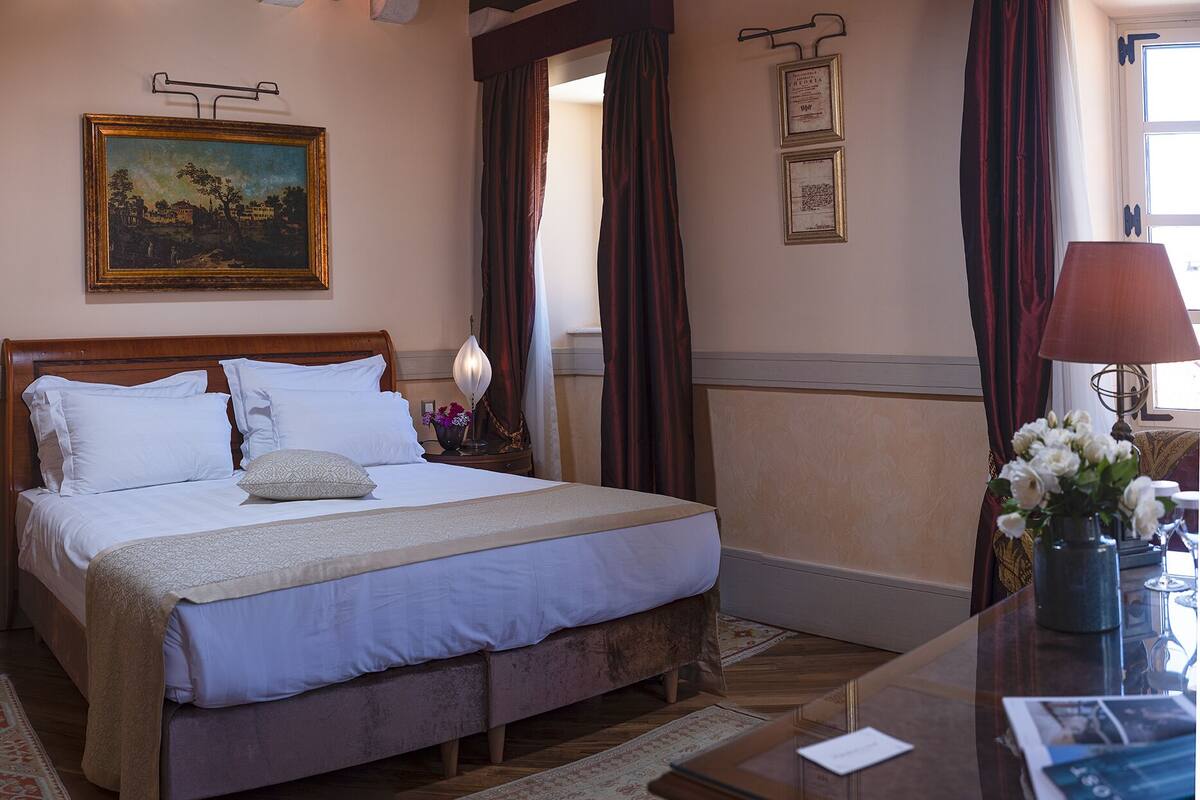 A hotel room with a large bed, a nightstand with a lamp, a painting on the wall, and a desk with a vase of flowers.
