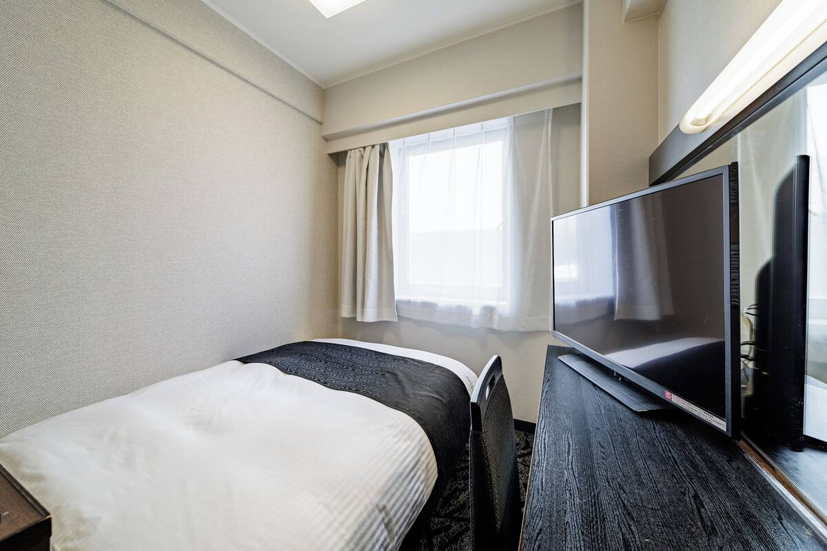 A hotel room with a bed, a desk, a flat-screen TV, and a window with curtains.