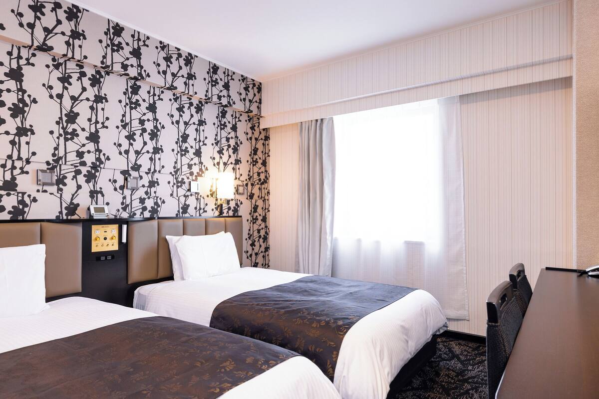 A hotel room with two beds, a desk, a control panel, and a large window with curtains.