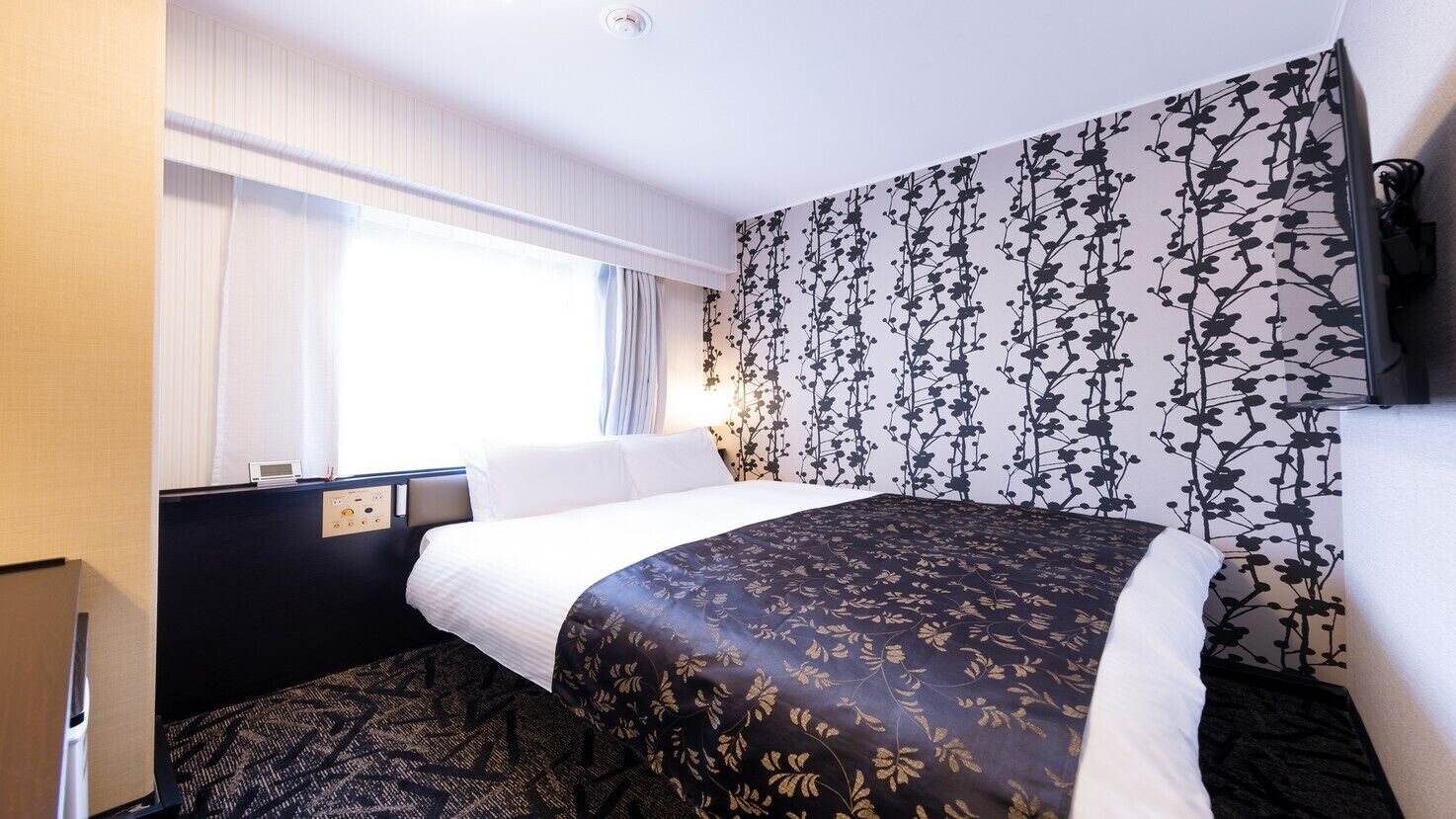 Une chambre d’hôtel avec un lit, un panneau de commande, une fenêtre avec des rideaux et un mur orné d’un motif floral.