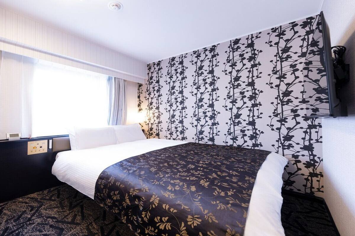 A hotel room with a bed, a control panel, a window with curtains, and a wall with a floral pattern.