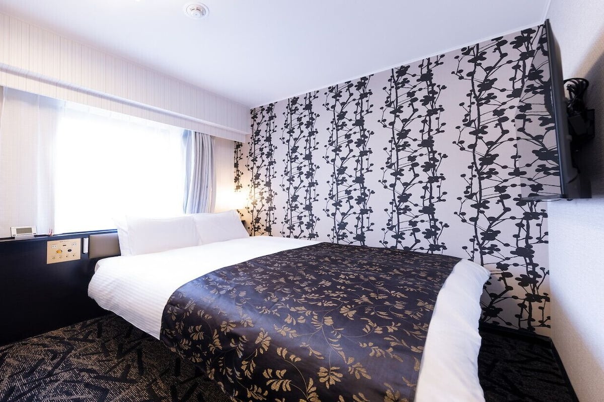 Ein Hotelzimmer mit Bett, Bedienfeld, Fenster mit Vorhängen und Wand mit Blumenmuster.