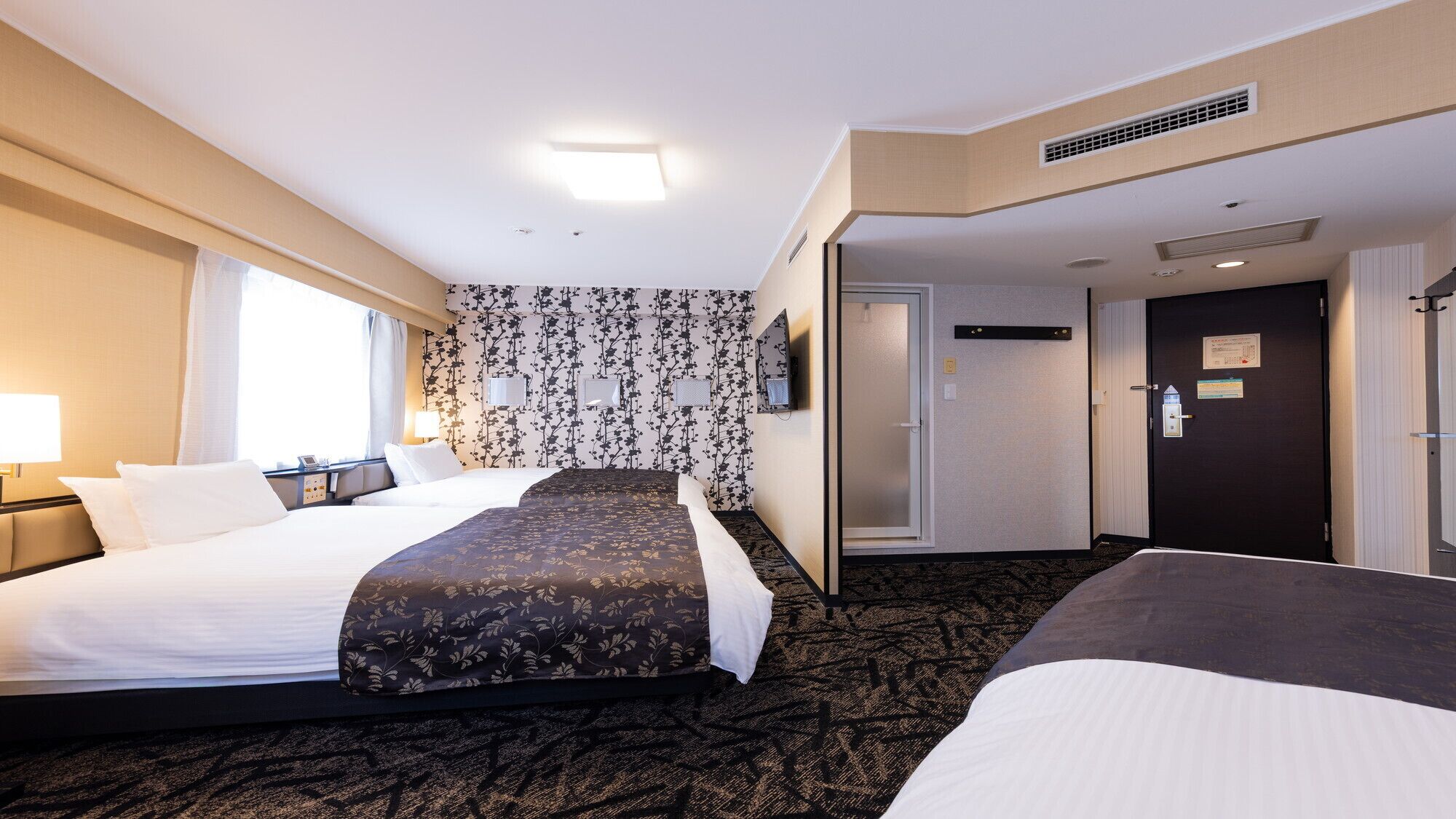 Une chambre d’hôtel avec deux lits, une grande fenêtre, un téléviseur fixé au mur et une porte donnant sur une autre pièce.