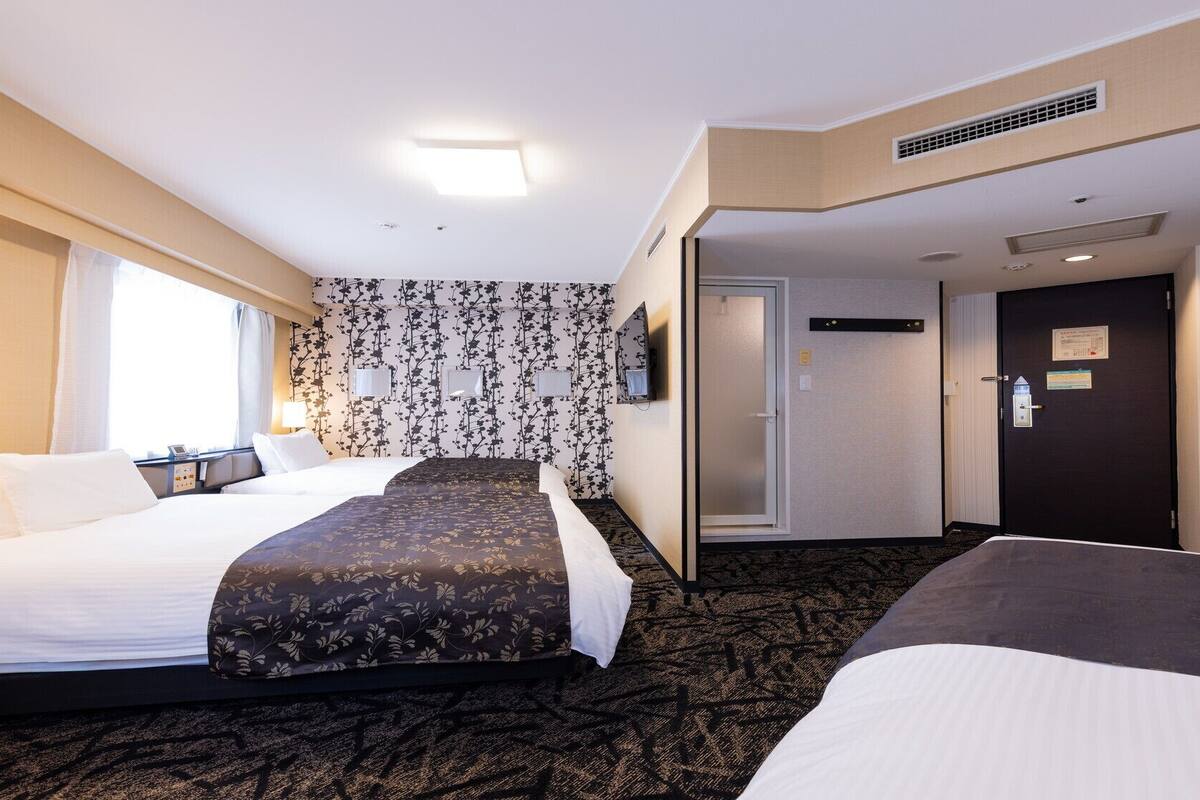 A hotel room with two beds, a large window, a wall-mounted TV, and a door leading to another room.