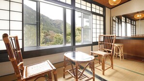 A room with tatami flooring, a low table, two chairs, and large windows offering a view of a mountainous landscape. - The Aizuya (Nasushiobara)