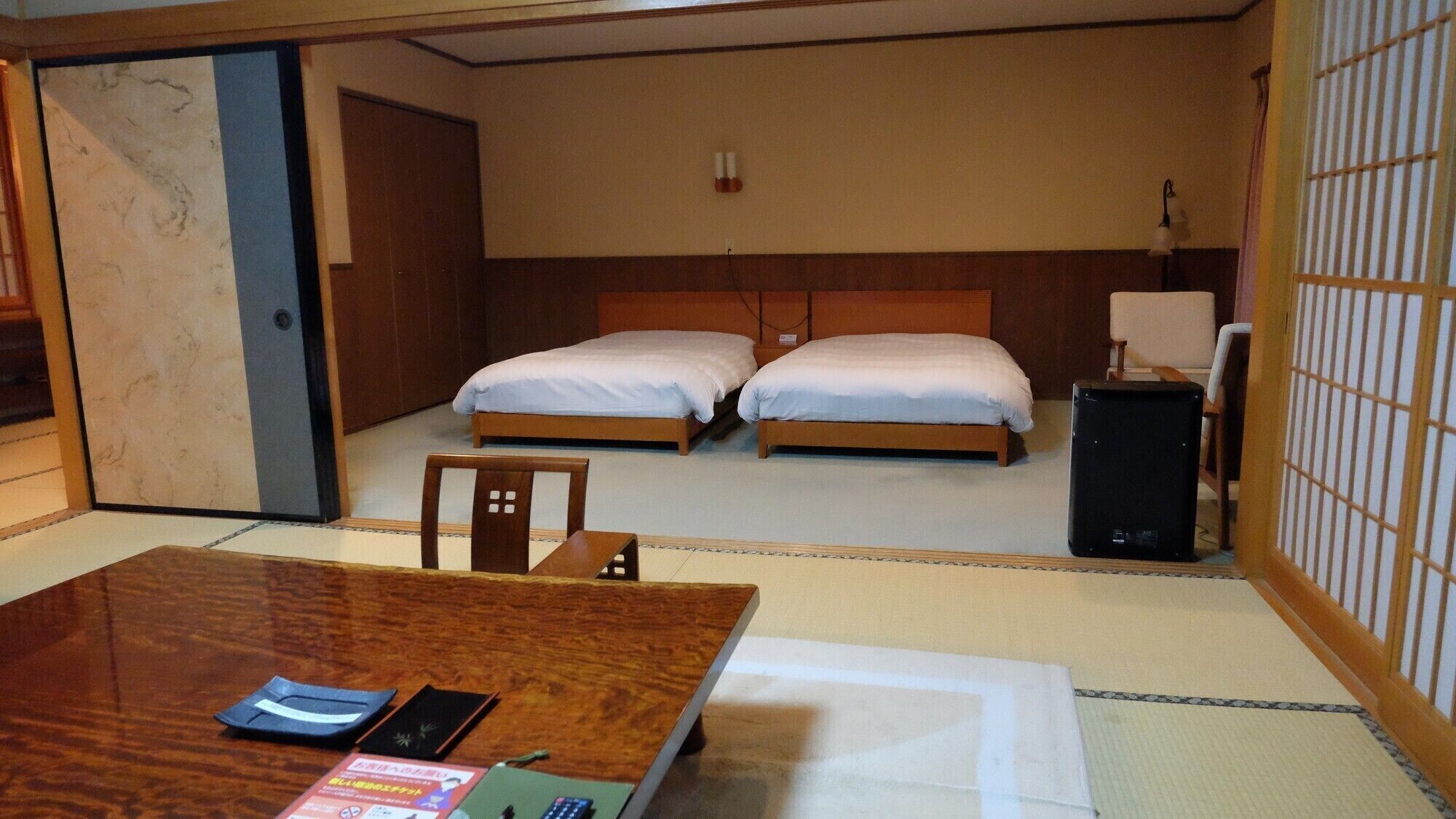 A hotel room with two beds, a desk with a chair, a bedside table, a lamp, and a traditional Japanese sliding door.