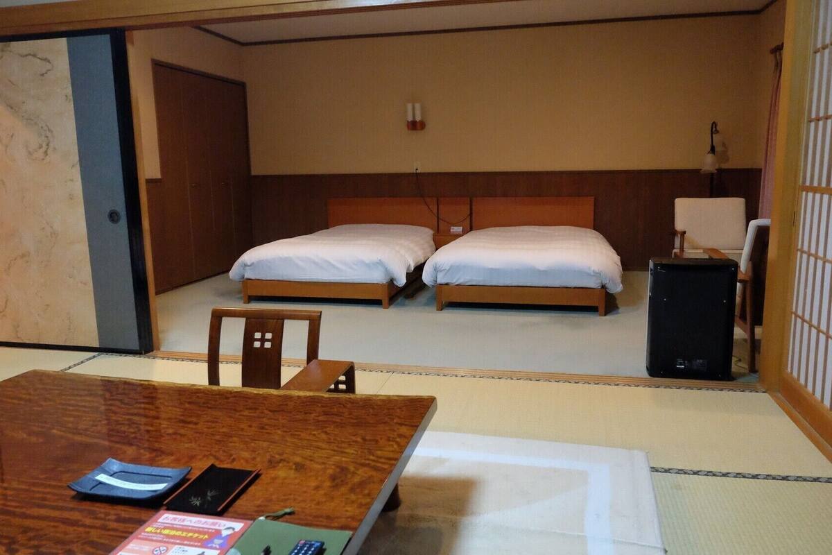 A hotel room with two beds, a desk with a chair, a bedside table, a lamp, and a traditional Japanese sliding door.