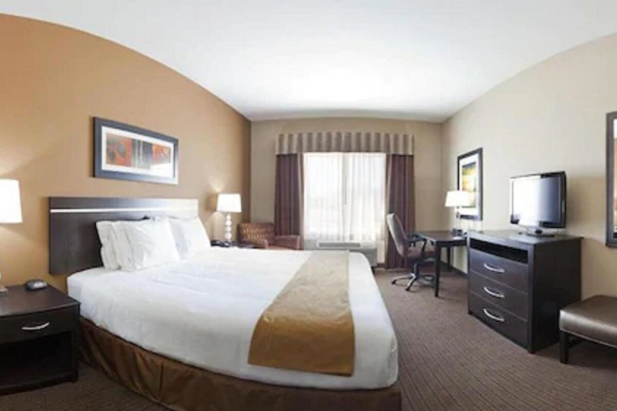 A hotel room with a large bed, two bedside tables with lamps, a desk with a chair, a television, and a window with curtains.