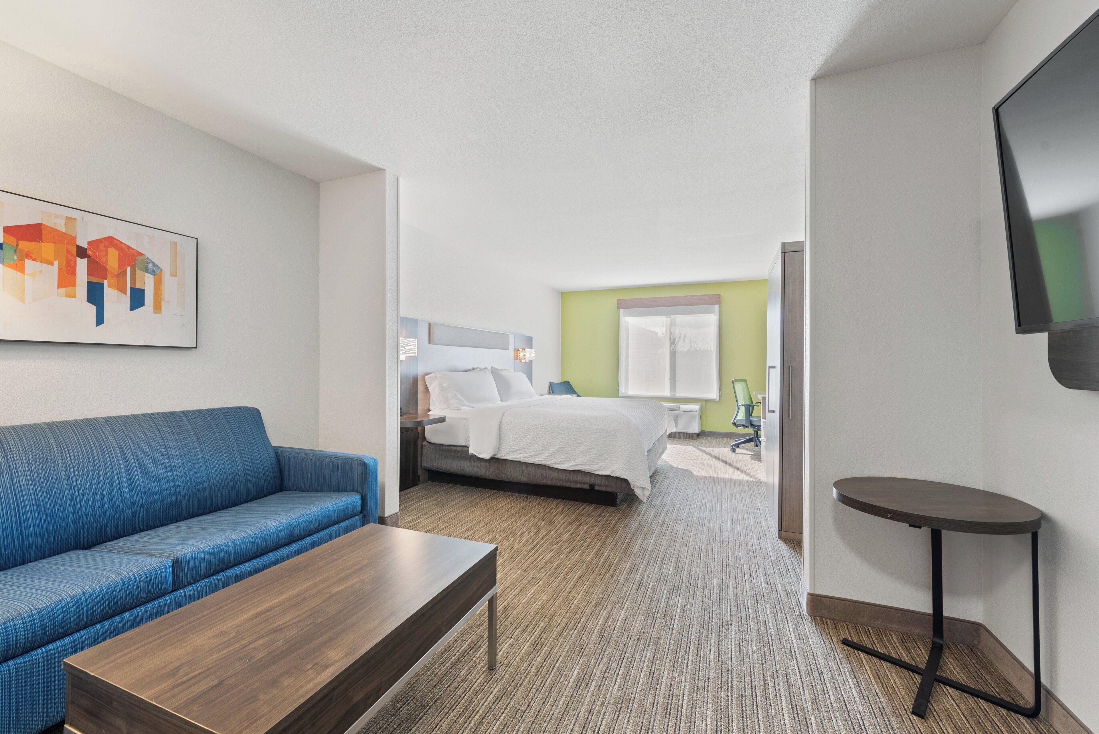 Una camera d'albergo con un divano blu, un tavolino in legno, un letto con biancheria bianca, una televisione a parete e un pavimento rivestito di moquette.