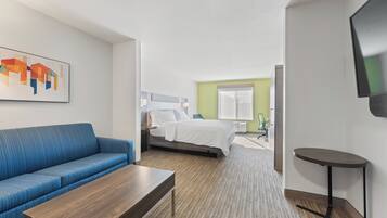 Una camera d'albergo con un divano blu, un tavolino in legno, un letto con biancheria bianca, una televisione a parete e un pavimento rivestito di moquette.