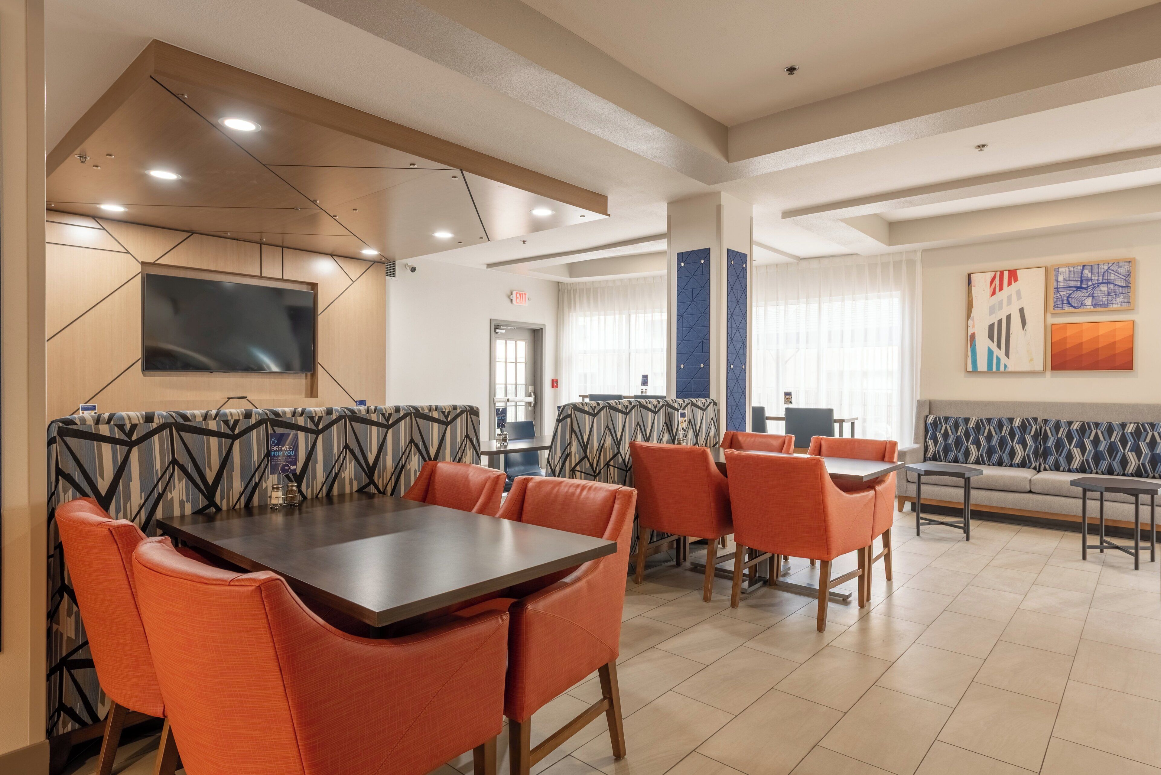 A modern dining area with a large table and orange chairs, a flat-screen TV mounted on the wall, and a seating area with patterned cushions.