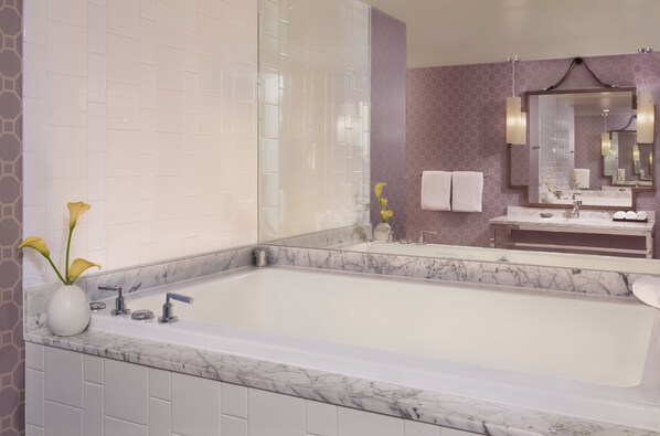 A modern bathroom with a large bathtub, a marble countertop, a white vase with flowers, and a large mirror with a purple wall.
