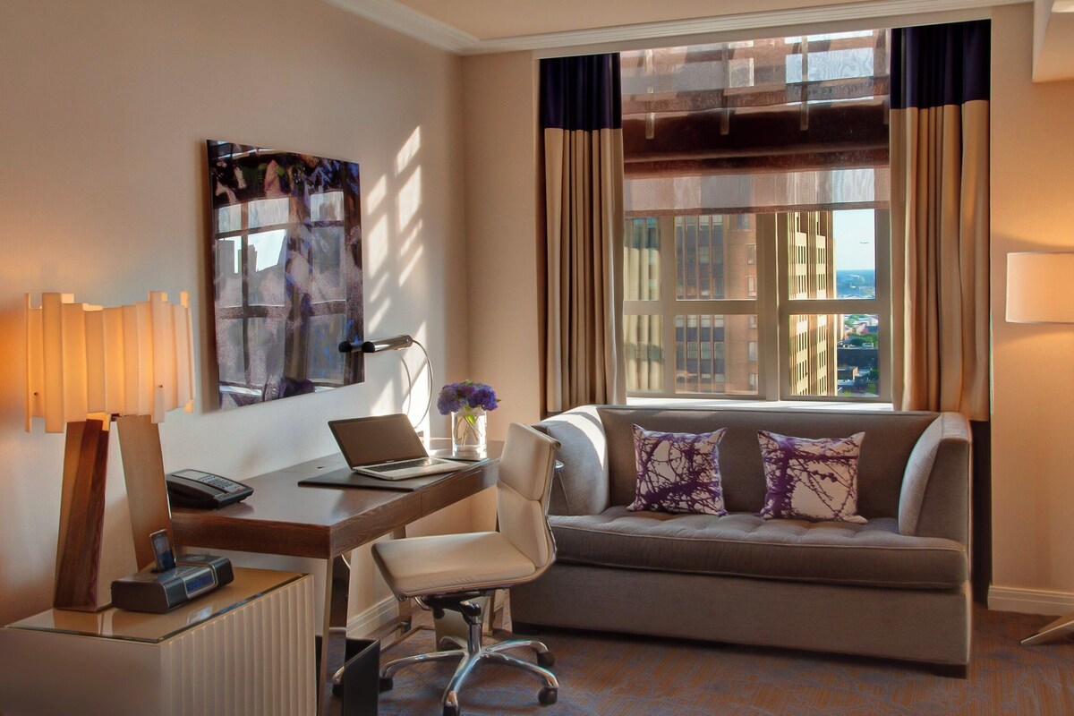 A hotel room with a desk, chair, sofa, and a large window with curtains.
