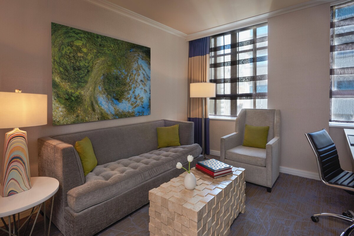 A living room with a grey sofa, a white coffee table, a side table, a lamp, a chair, and a large abstract painting on the wall.