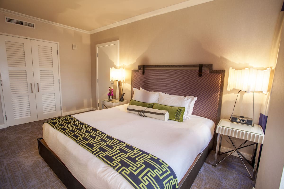 Un letto rifatto con cura, una coperta a motivi decorativi, un comodino con una lampada e uno specchio fissato alla parete.