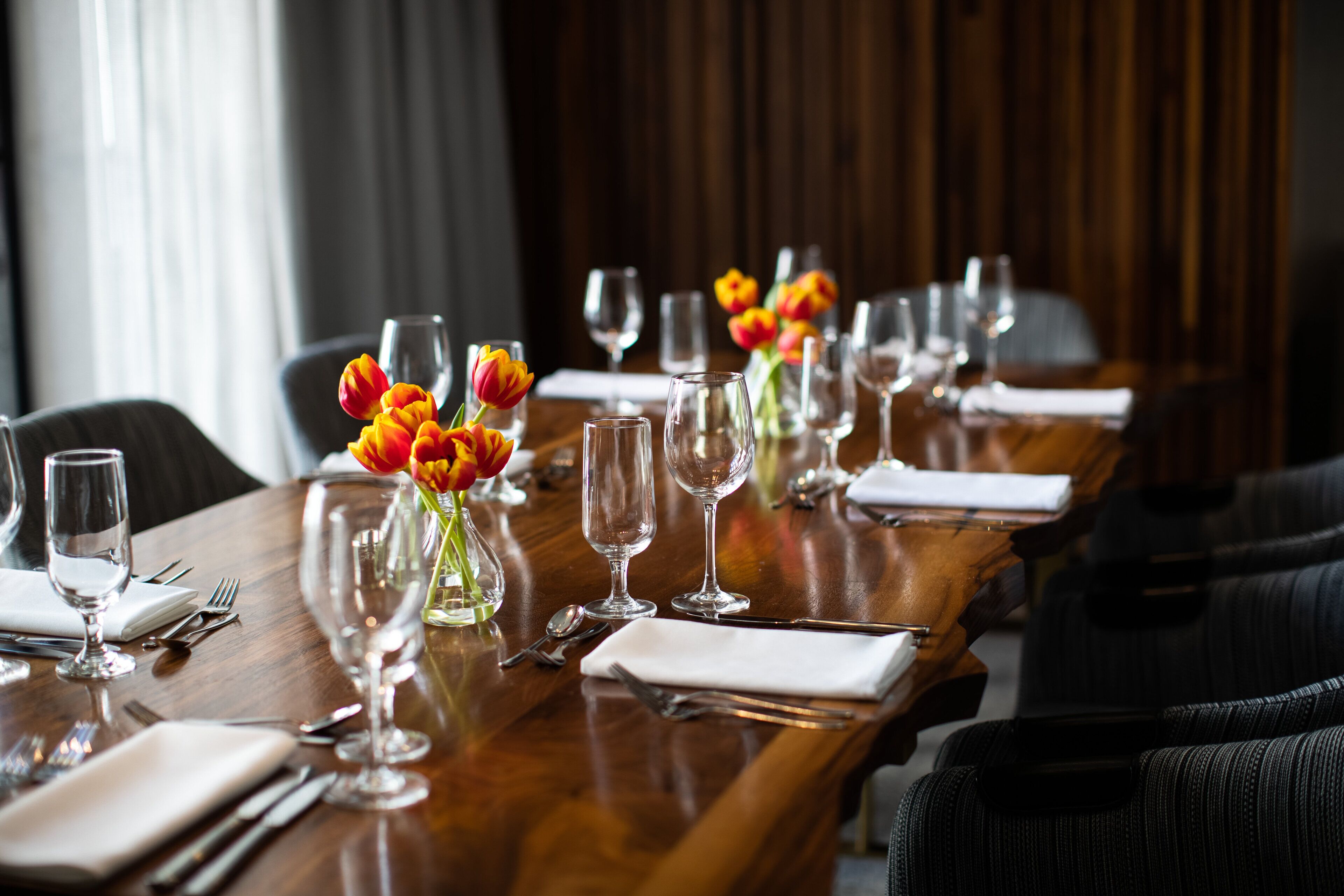 A long wooden table set for dining with multiple wine glasses, a vase with flowers, and neatly arranged cutlery.
