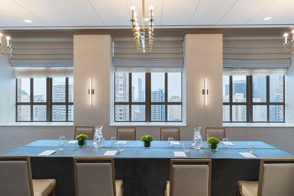 A conference room with a long table, chairs, and large windows offering a city view.