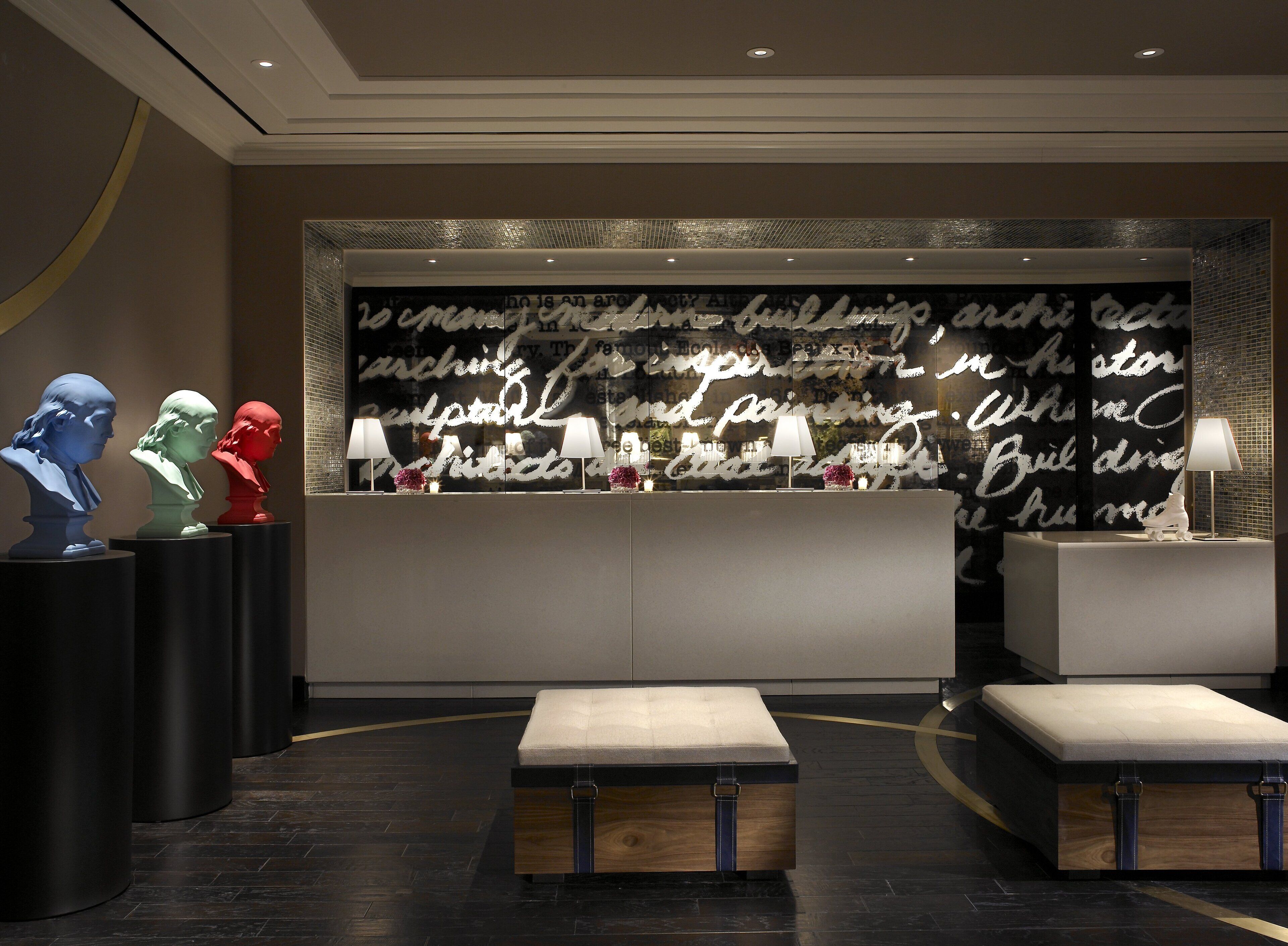 A reception area with a modern design, featuring a large wall with artistic text, three sculptural busts, and two illuminated lamps.