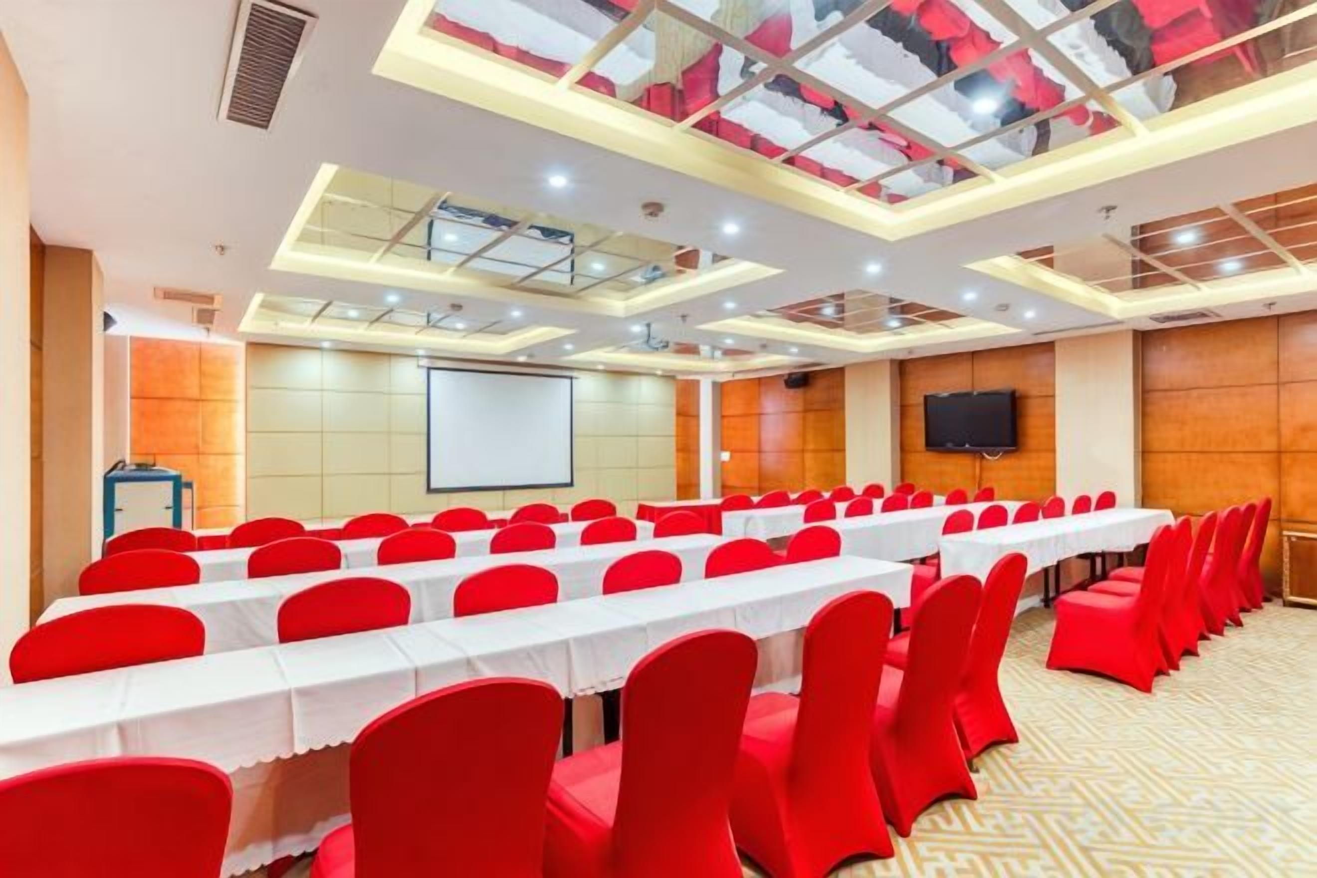Un salón de conferencias con una mesa larga, sillas rojas, una pantalla de proyector y un televisor en la pared.