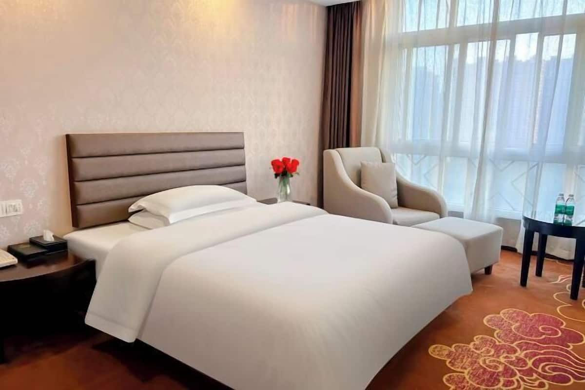 Habitación de hotel con una cama grande, una silla, una mesita y un florero con flores.