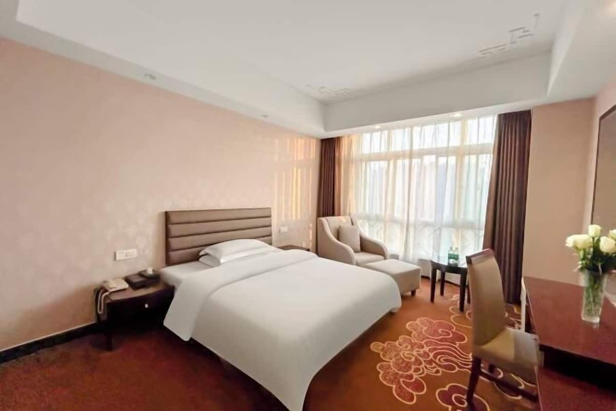Una habitación de hotel con una cama grande, una zona de estar con una silla, una mesita y un ventanal con cortinas.