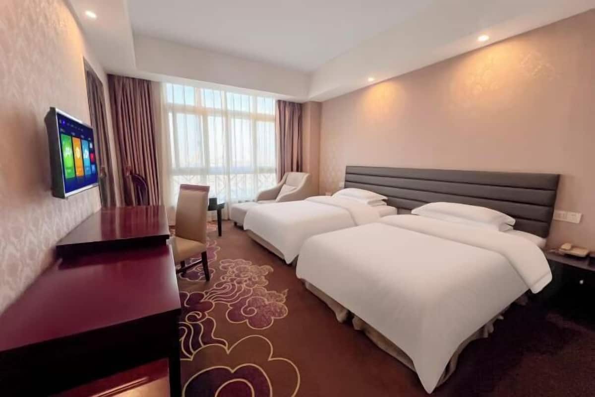 Habitación de hotel con dos camas, un escritorio, un televisor y un ventanal grande con cortinas.