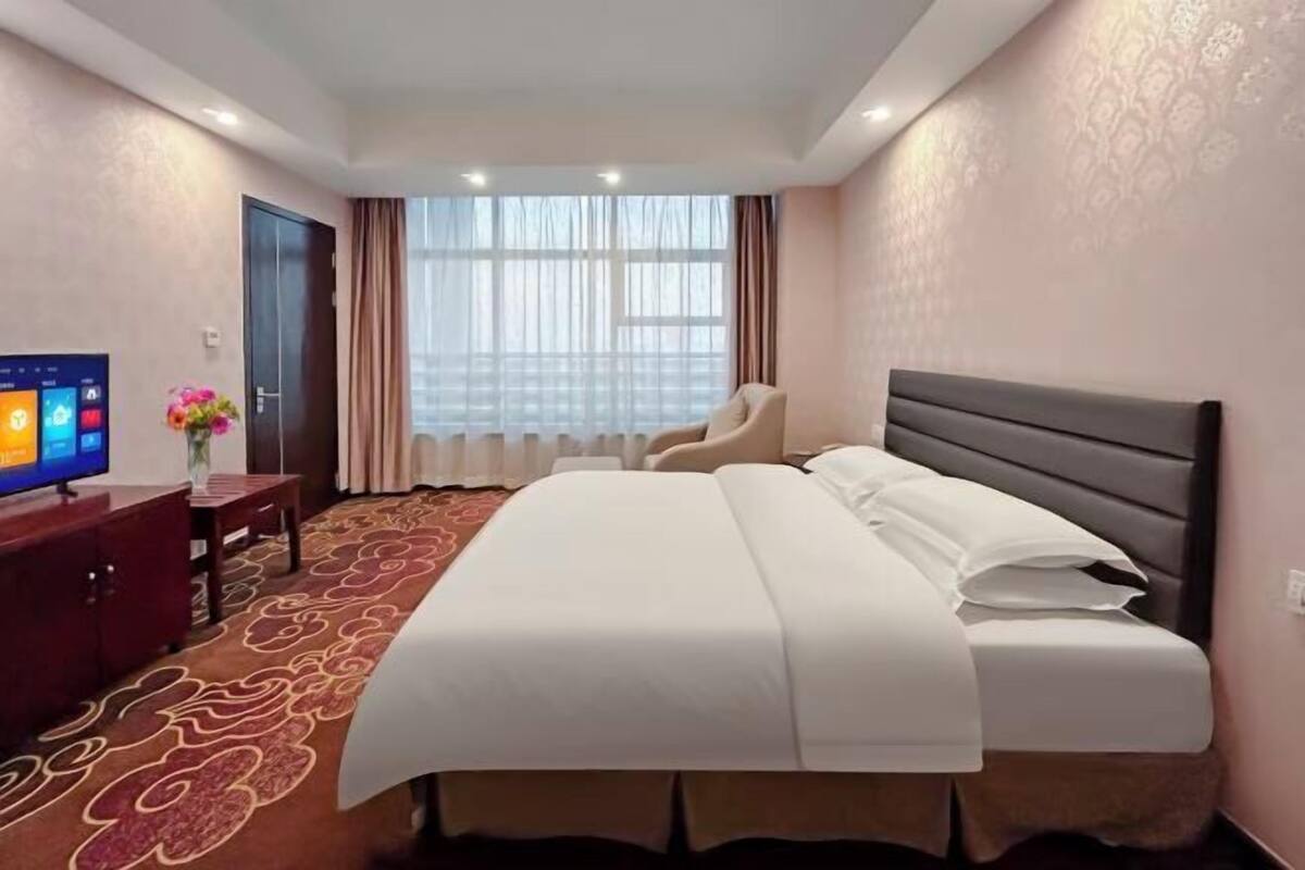 Habitación de hotel con una cama grande, televisor, escritorio con un florero y ventana con cortinas.
