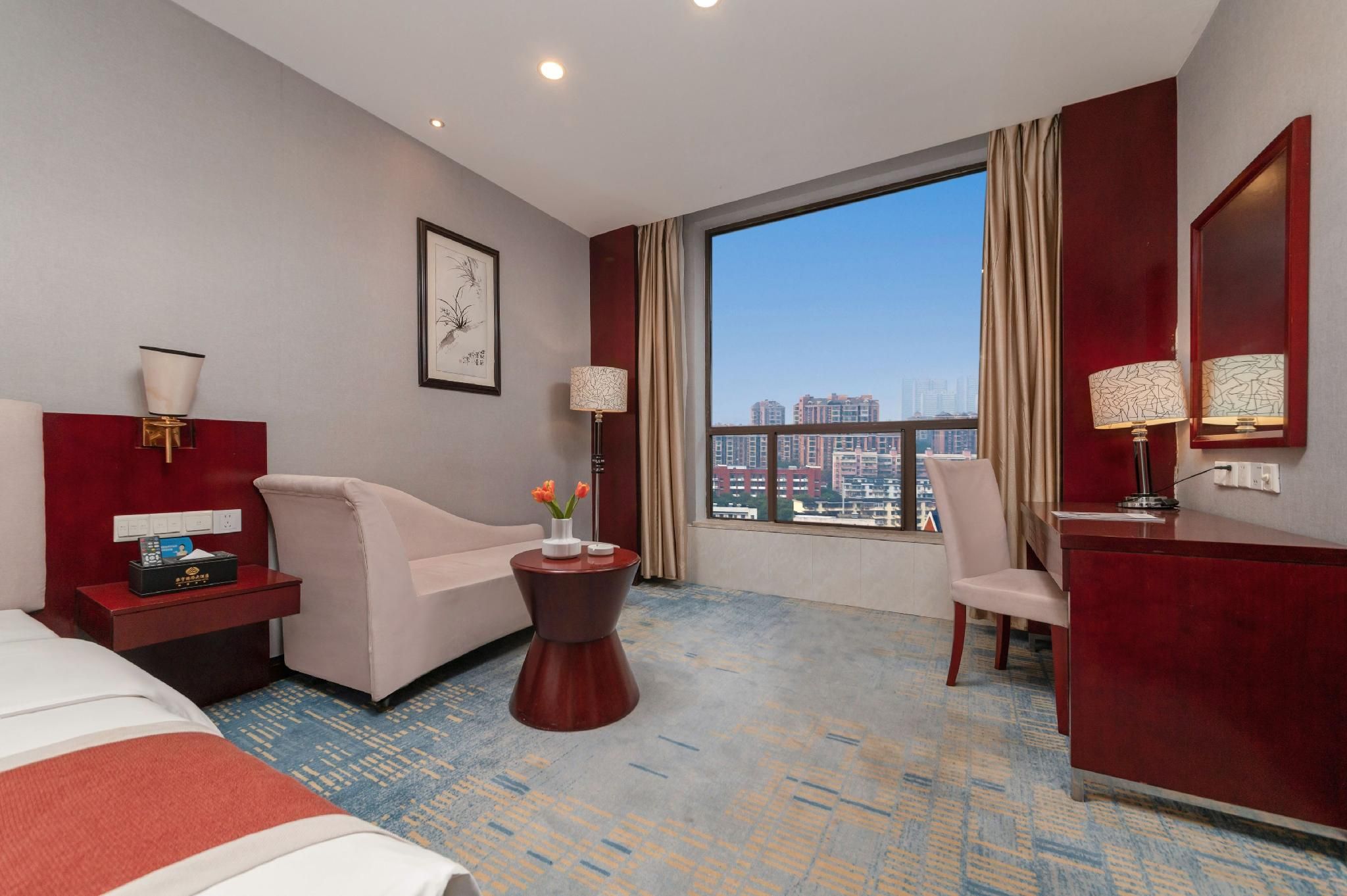 A hotel room with a bed, a sofa, a desk, a chair, a small table with flowers, a lamp, a painting, and a large window with a city view.