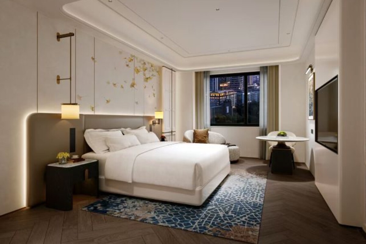 A modern hotel room with a large bed, a small table, a TV, and a city view.