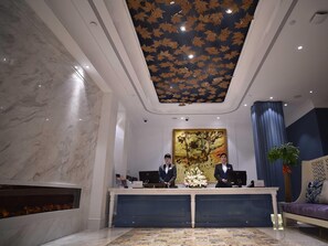 A hotel reception area with marble walls, a ceiling with leaf patterns, a large floral painting, and a reception desk with two staff members. - Jinchen Hotel (Shanghai)