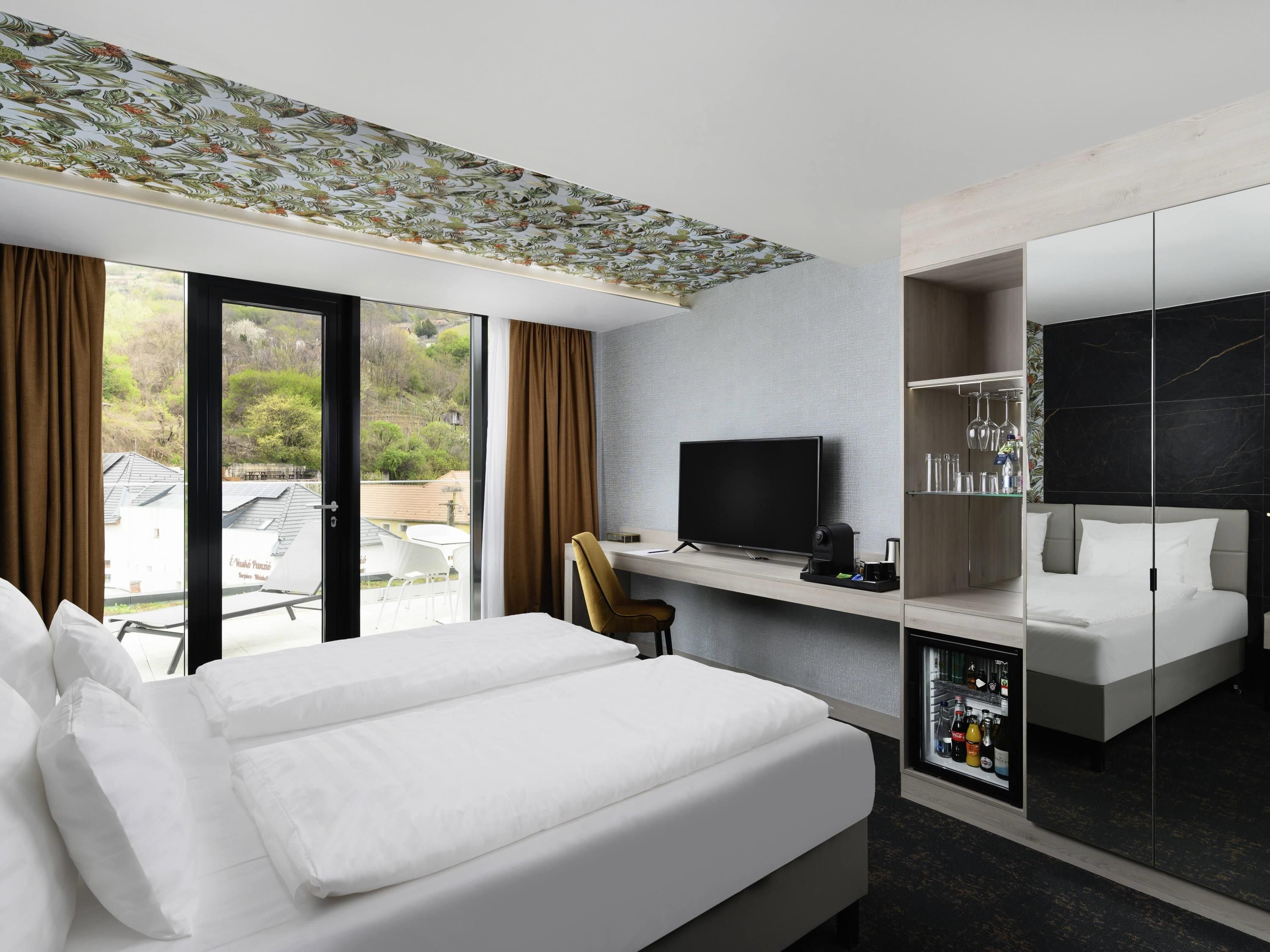A hotel room with a bed, a desk with a TV, a minibar, and a large window with curtains.