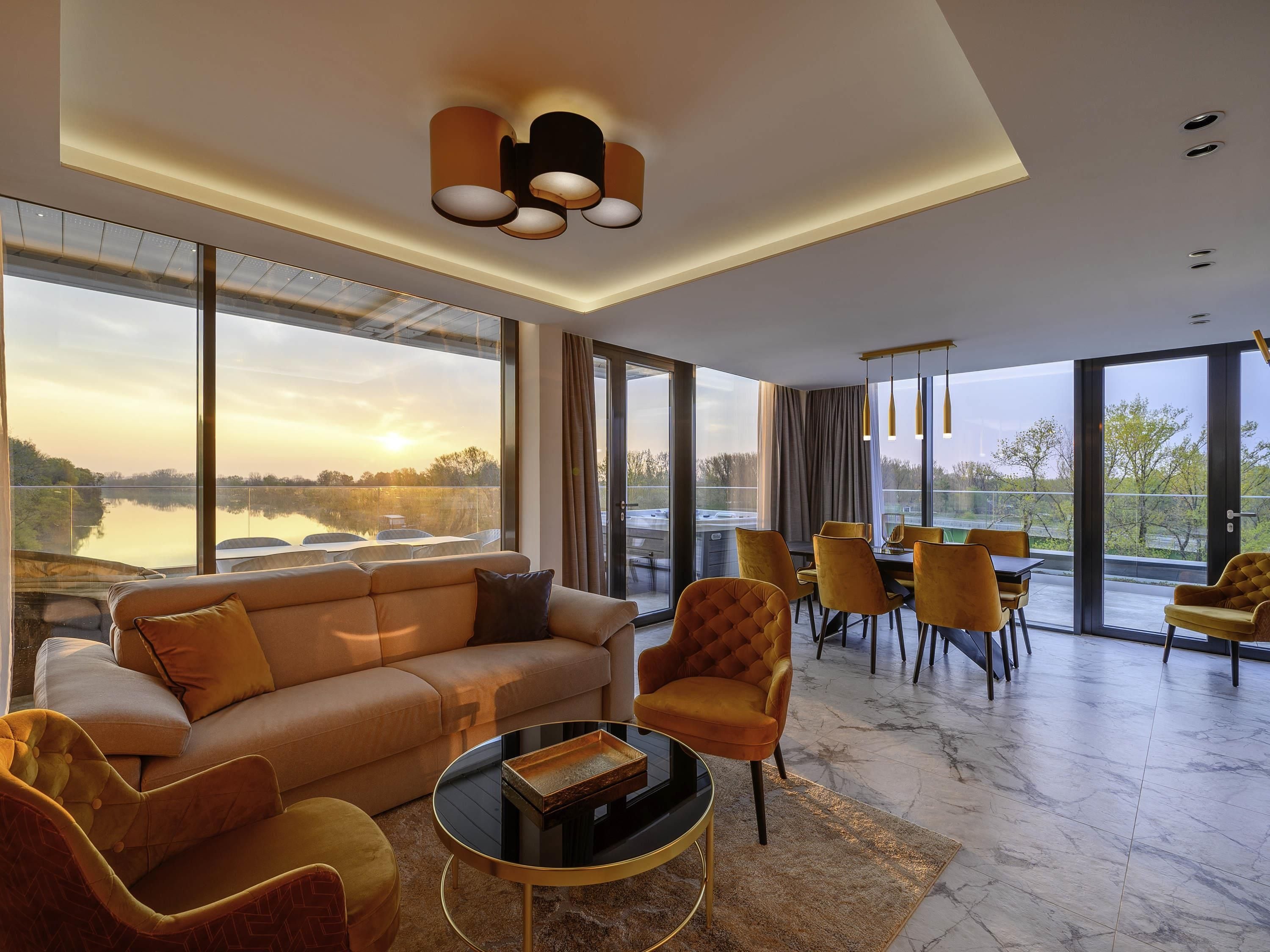 Uma sala de estar moderna com um sofá grande, poltronas, uma mesa de centro e uma área de jantar com cadeiras e uma mesa. Há janelas amplas com vista para um corpo de água e árvores.