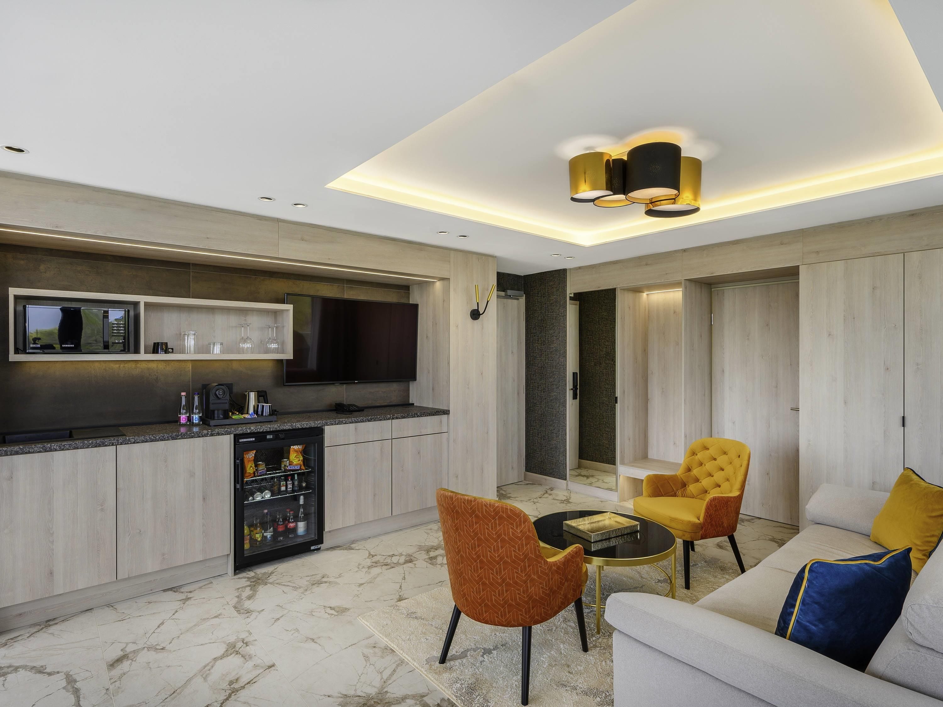A modern kitchen with a built-in microwave, a refrigerator, and a flat-screen TV. There is a seating area with a sofa and armchairs, and a small round table.