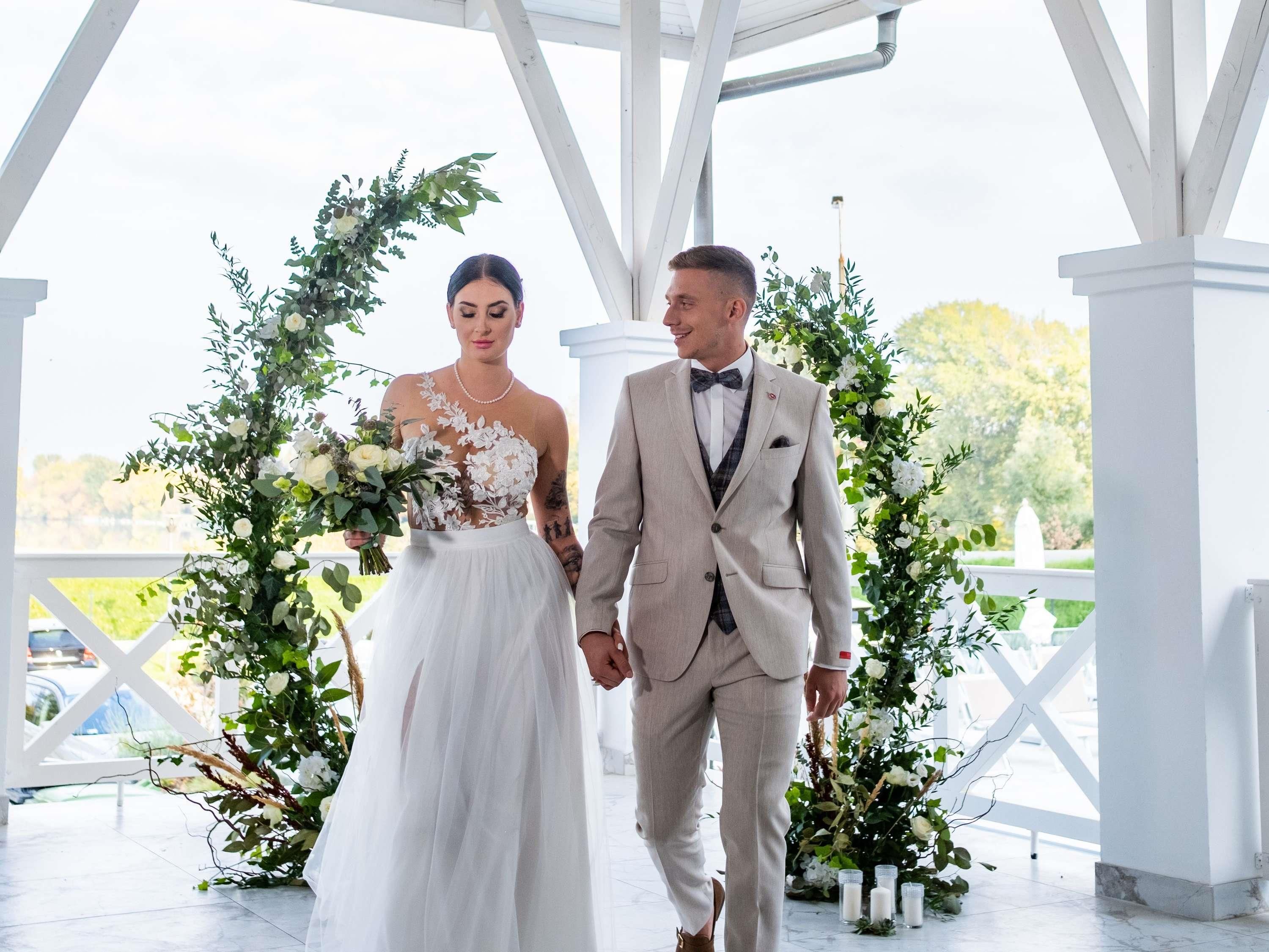 A couple holding hands in a covered outdoor area with white columns and floral arrangements.