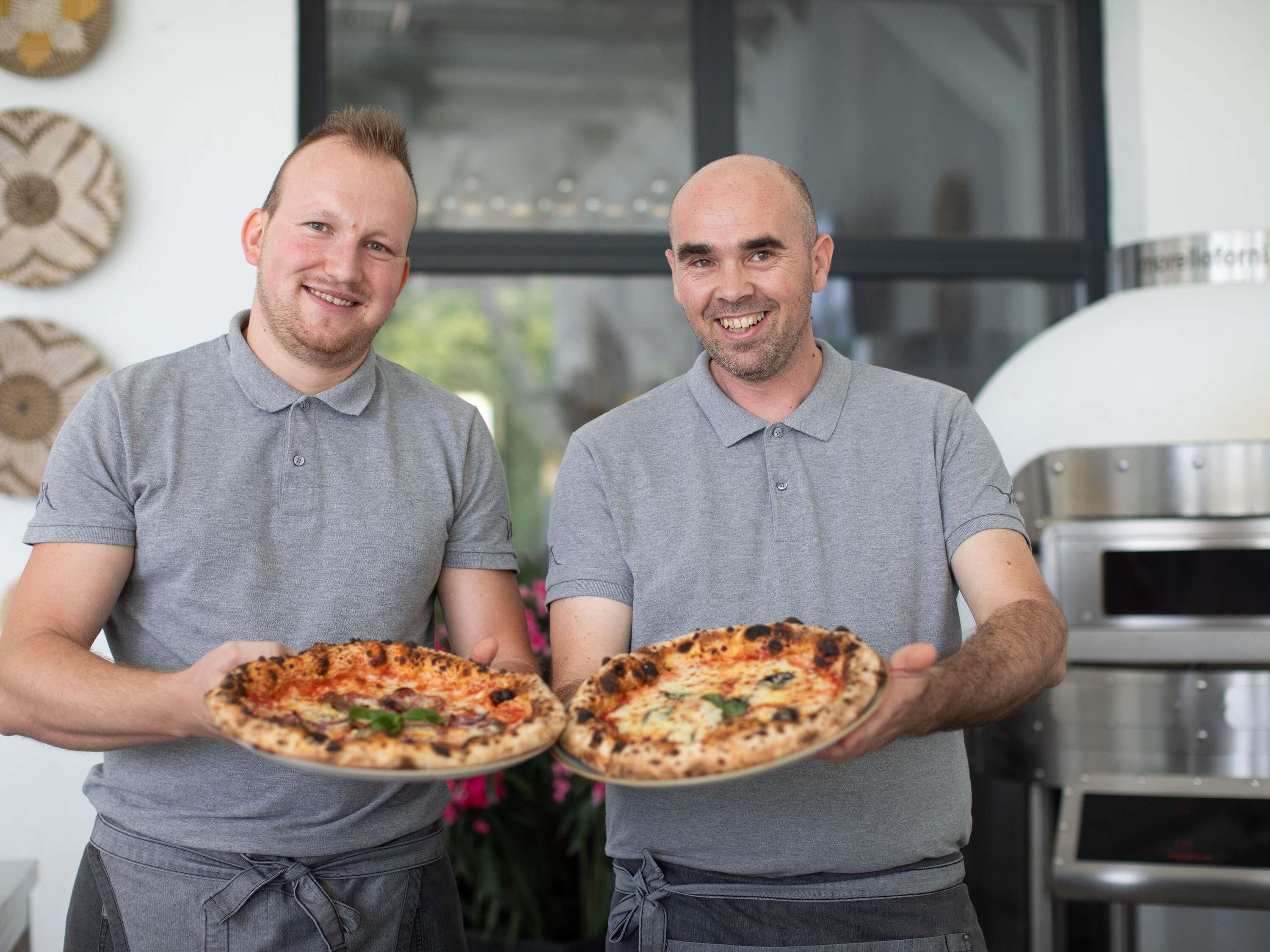 Dos personas sosteniendo pizzas en una cocina, con un horno de pizza al fondo.