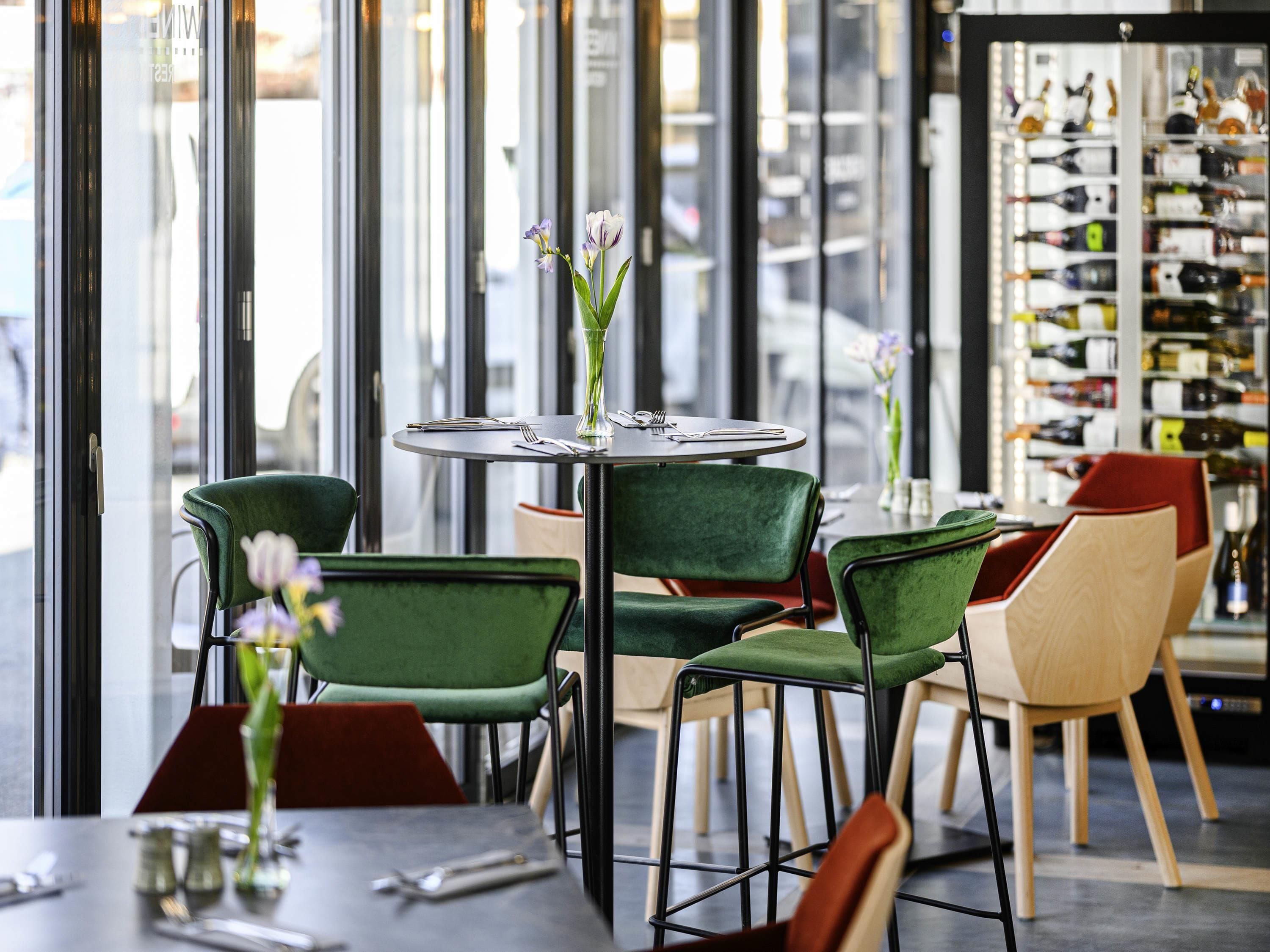 Un café moderno con mesas redondas, sillas verdes y rojas, y una exhibición de vinos al fondo.
