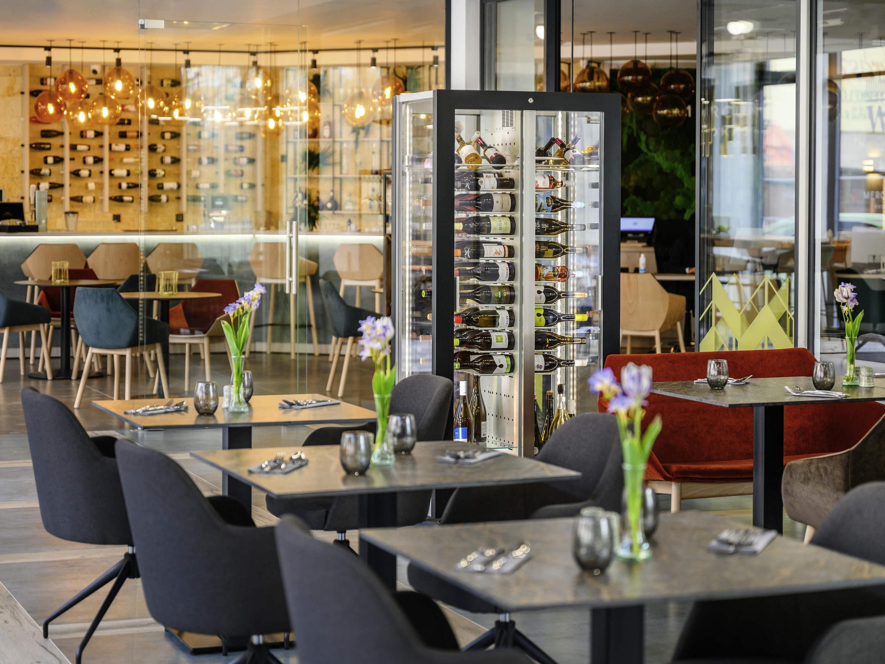 Interior de un restaurante con mesas, sillas, un refrigerador para vino y una pared llena de botellas.