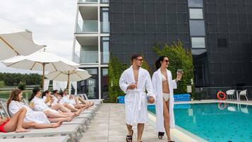 A poolside area with lounge chairs and umbrellas, a modern building with balconies, and a swimming pool.