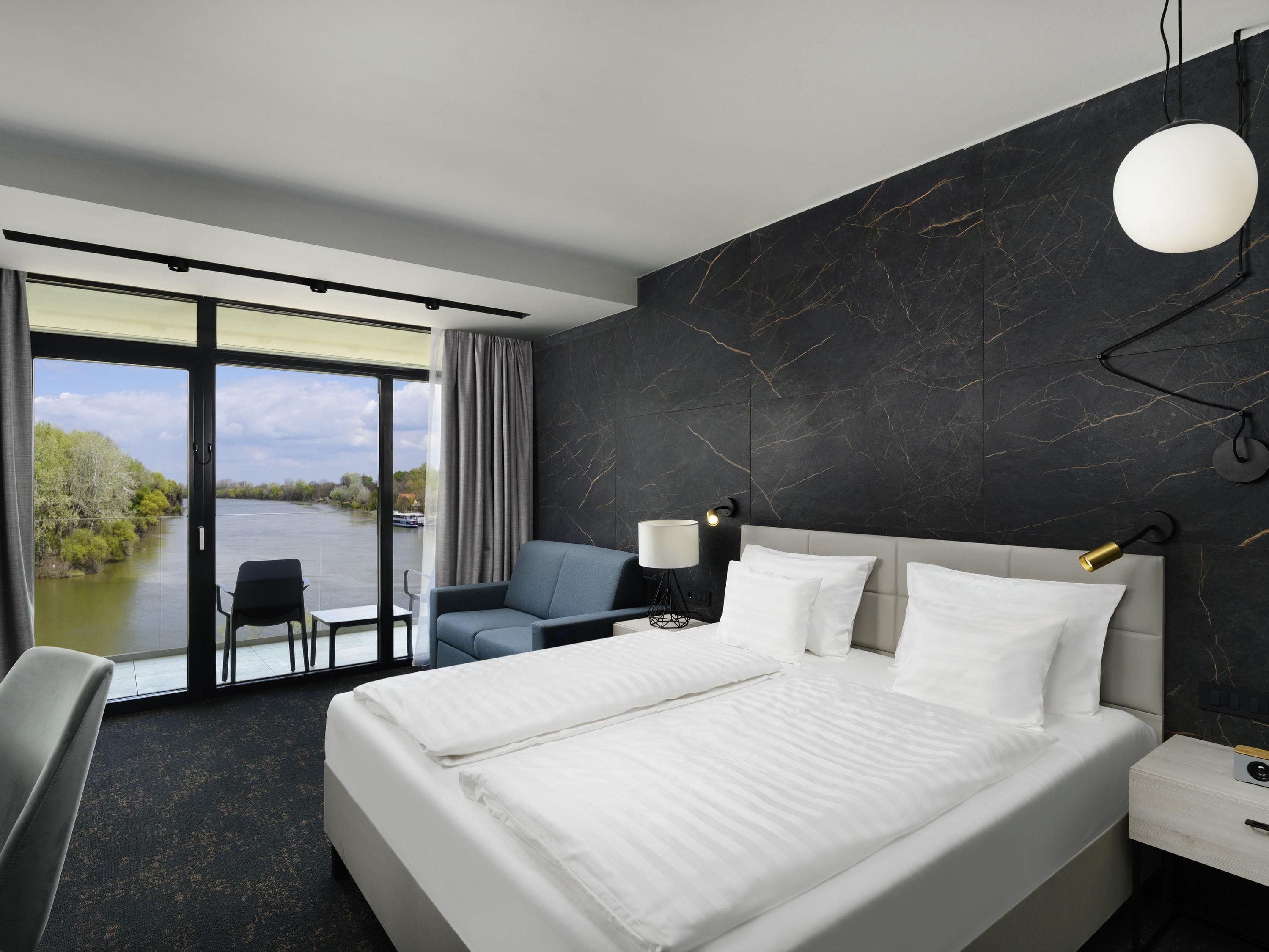 Een moderne hotelkamer met een groot bed, een bureau met stoel, uitzicht op een rivier en een donkere marmeren wand.