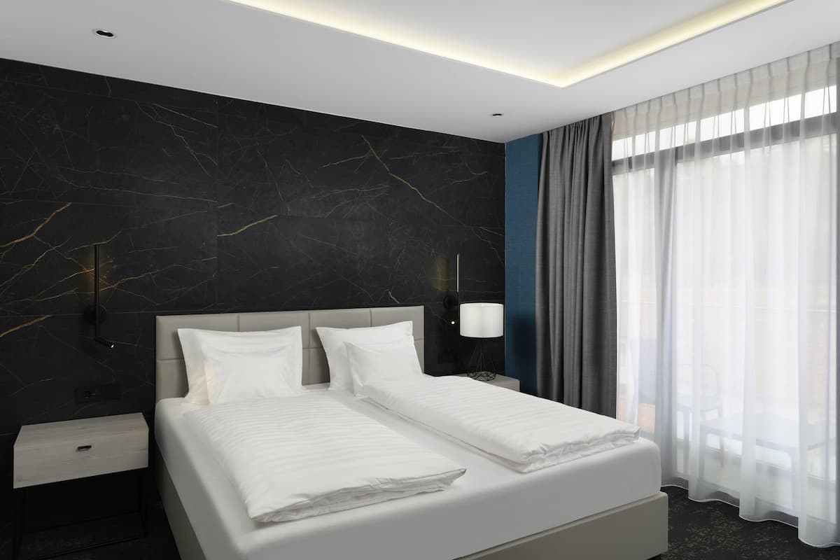 A modern hotel room with a large bed, a nightstand, a wall-mounted lamp, and a window with sheer curtains.