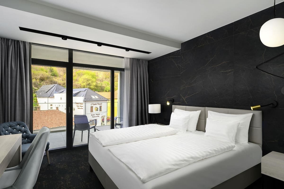 A modern hotel room with a large bed, a desk, and a view of a building through a sliding door.