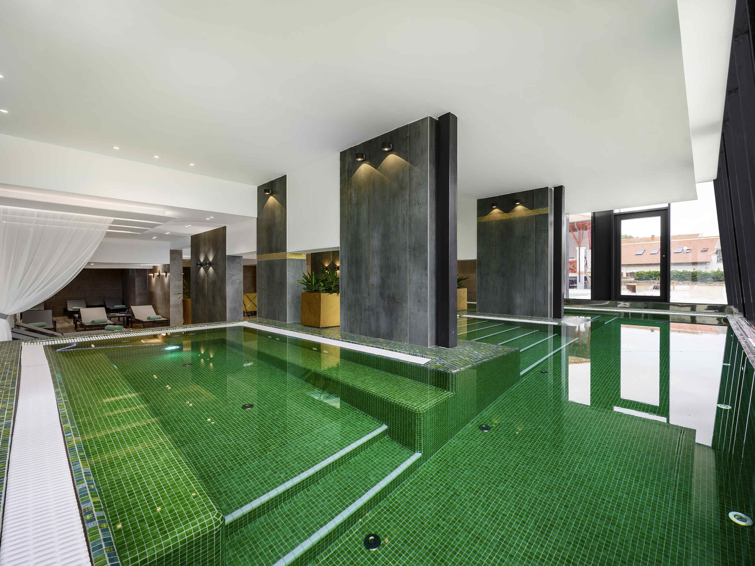 Uma piscina interior moderna, com chão em azulejos verdes, espreguiçadeiras e pilares grandes.
