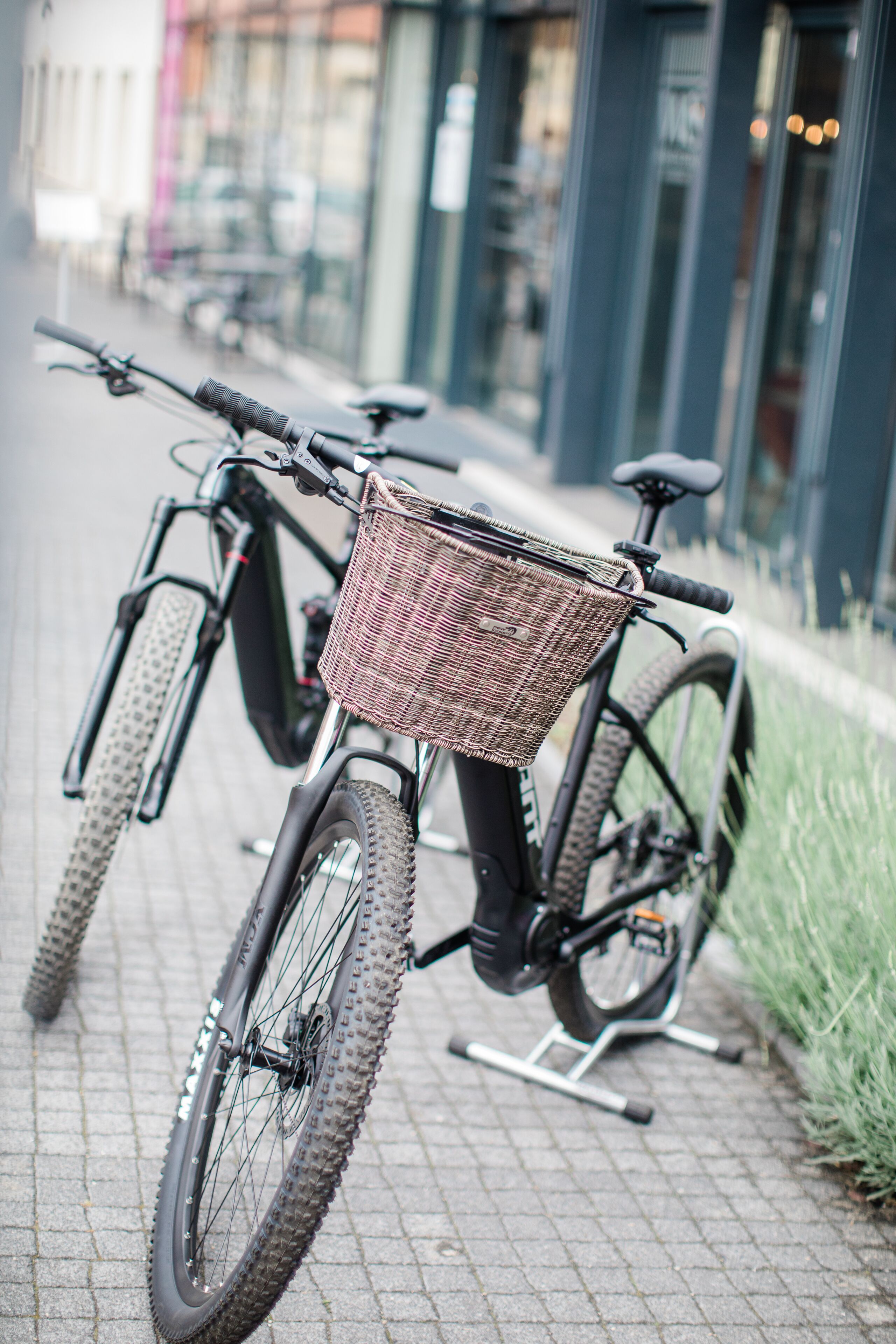 A bicycle with a wicker basket attached to the front, parked on a paved sidewalk.