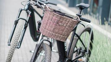 A bicycle with a wicker basket attached to the front, parked on a paved sidewalk.